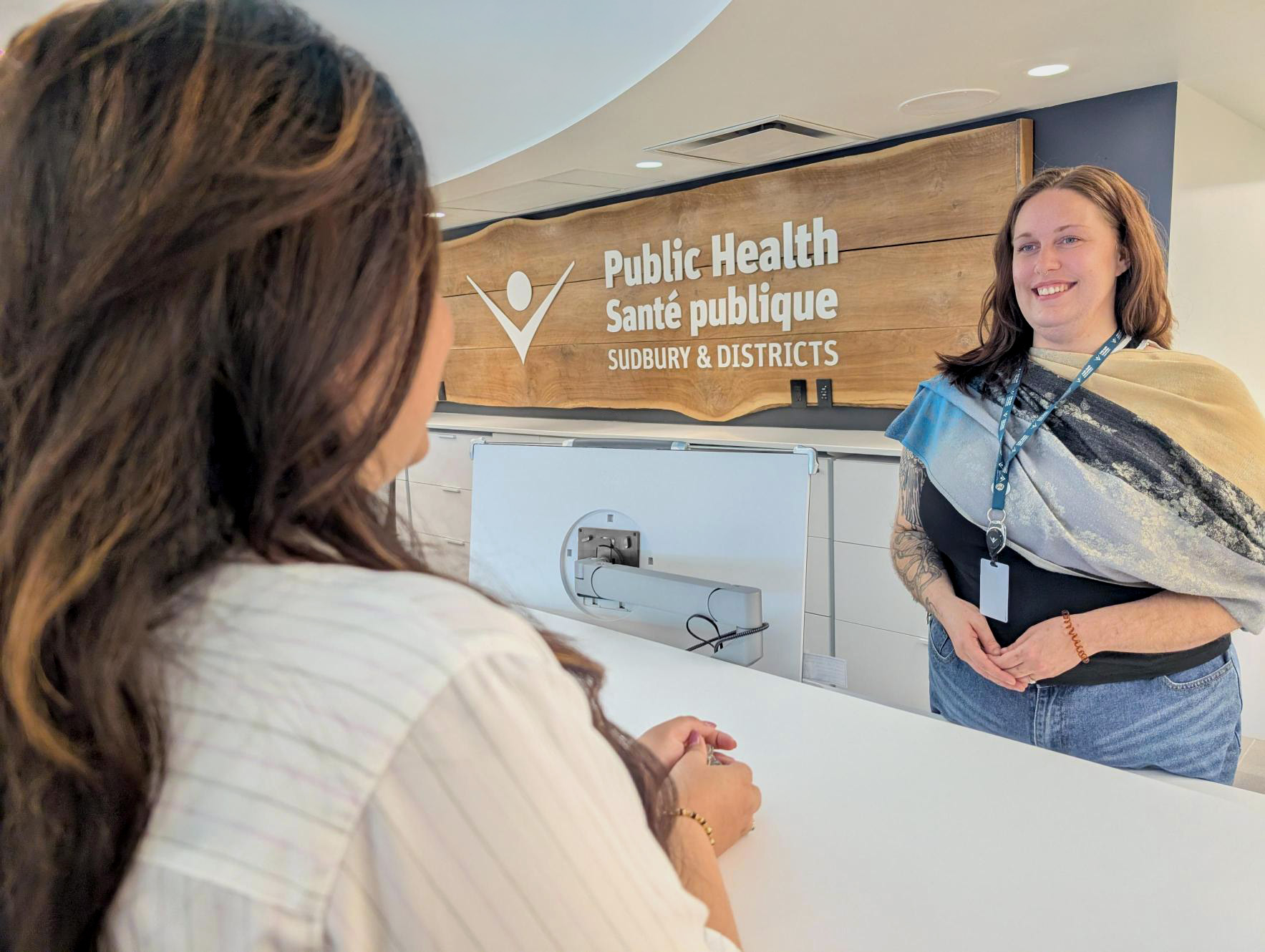 A photo of a Public Health Sudbury & Districts staff member greeting a client at the intake desk in front of the Public Health Sudbury & Districts logo.